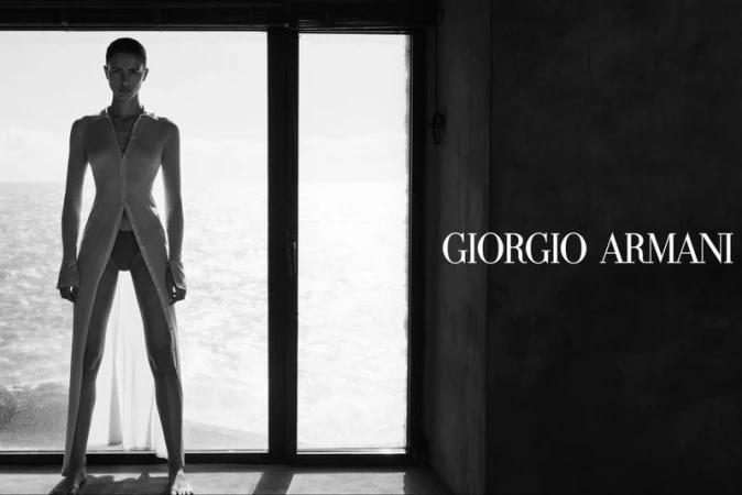 Black-and-white fashion shot of a woman in a translucent robe standing in front of a window, the ocean visible in the background.