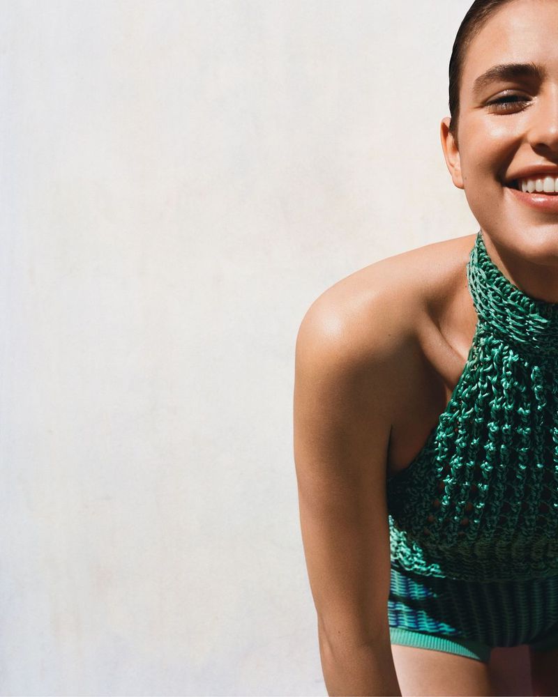 An adult female smiles in a medium shot with green halter top and striped shorts against a neutral background in outdoor light.