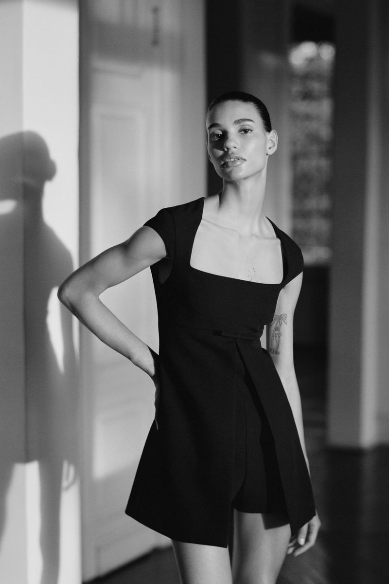 Black and white portrait of a woman in a dark dress, standing indoors with sunlight shadows casting on the wall and hardwood floor.
