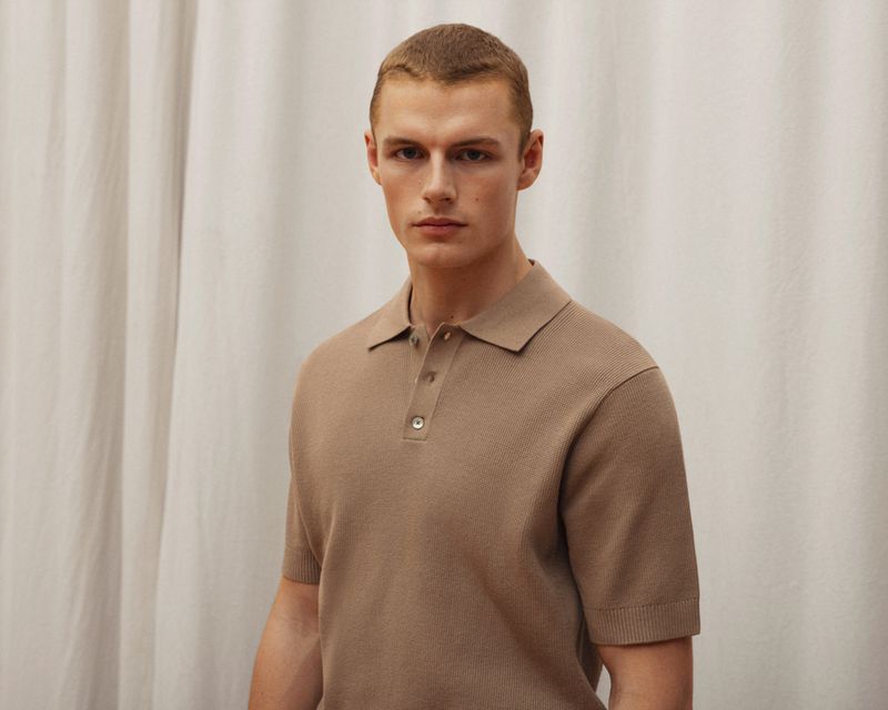 A fair-skinned adult male poses indoors wearing a short-sleeved, buttoned polo shirt against a neutral, draped textile background.