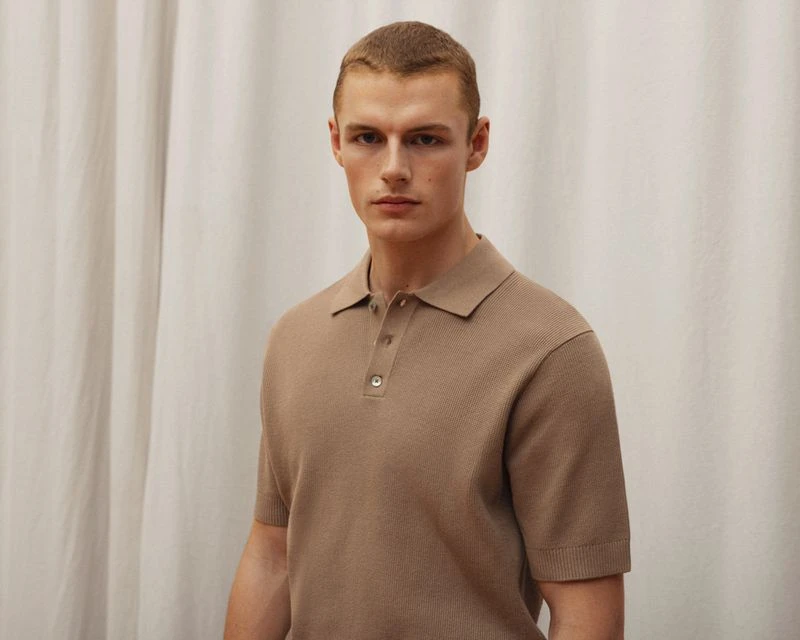 A fair-skinned adult male poses indoors wearing a short-sleeved, buttoned polo shirt against a neutral, draped textile background.