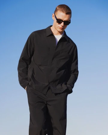 Adult man in a black collared shirt and black pants stands against a clear blue sky, wearing sunglasses in a medium studio shot.