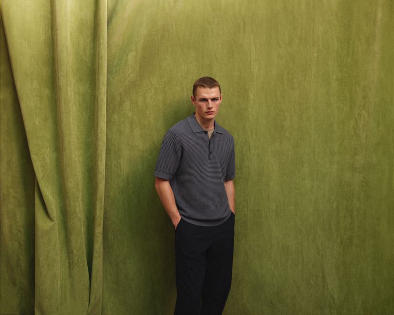 Portrait of a young adult man with short brown hair wearing a gray polo shirt and dark pants, posing in front of a green curtain backdrop.