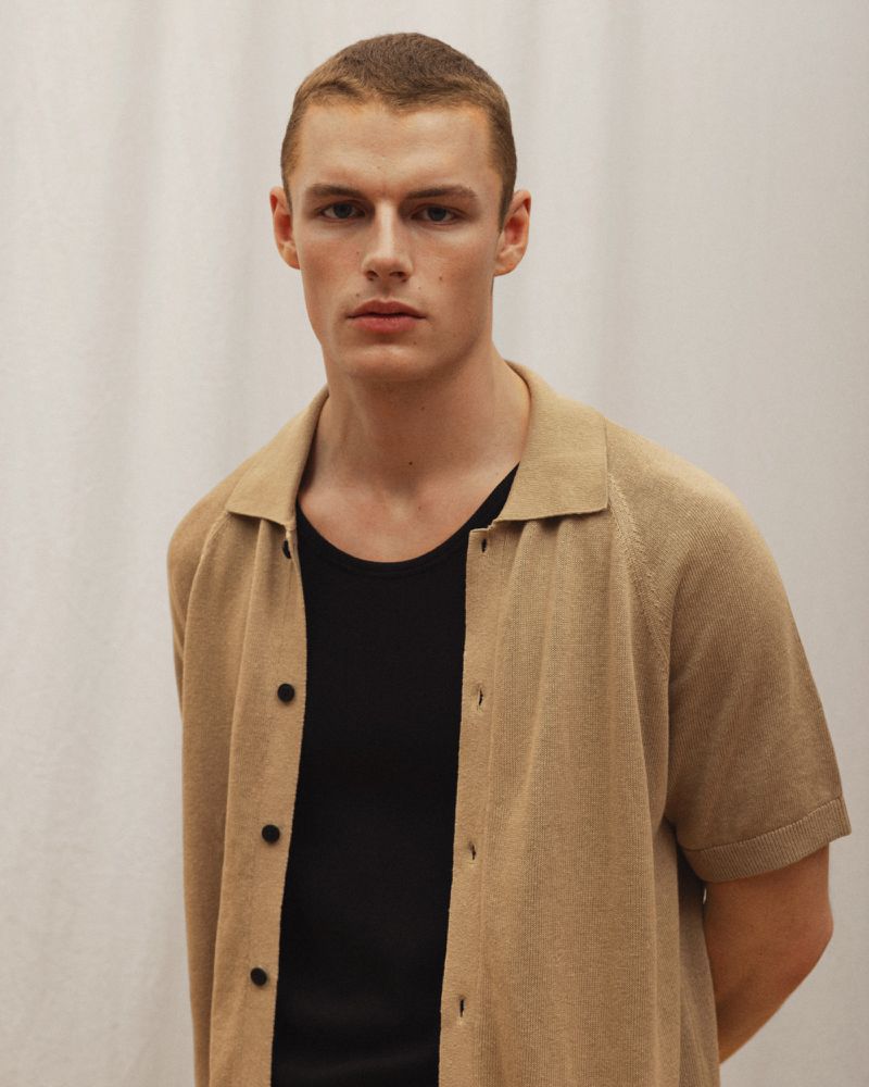 Man in casual attire: Close-up studio portrait of a light-skinned male wearing an open tan collared shirt over a black t-shirt.