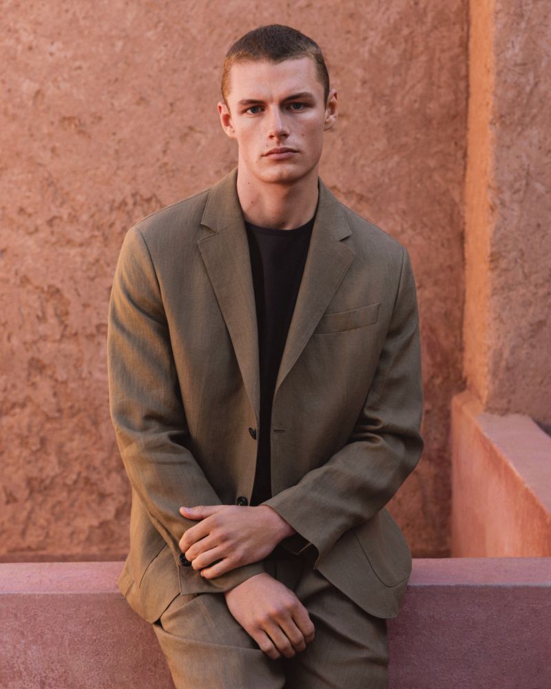 Portrait of a light-skinned male model in a green suit, posing outdoors with hands clasped against a textured orange wall backdrop.