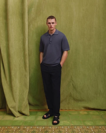 A casually dressed man in a blue polo shirt, black trousers, and black sandals poses against a textured green curtain backdrop.