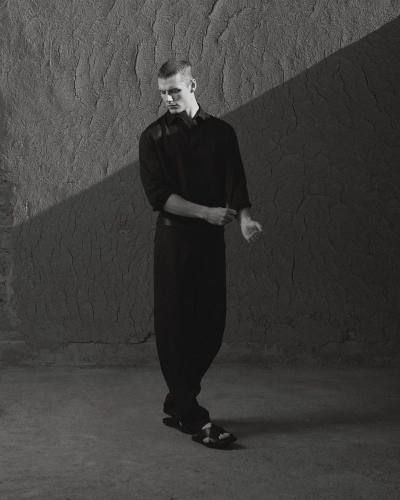 Black and white portrait of a young male fashion model posing against a textured wall, wearing a black shirt and pants.