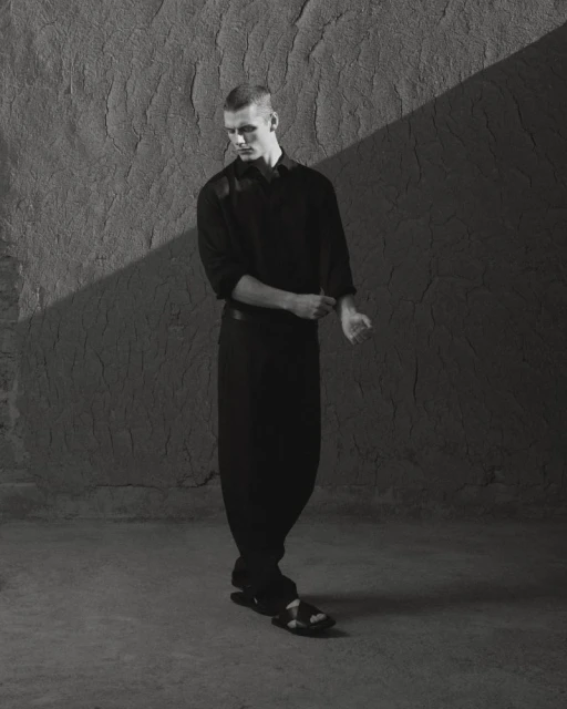 Black and white portrait of a young male fashion model posing against a textured wall, wearing a black shirt and pants.