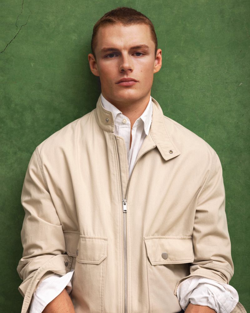 Close-up portrait of a male model wearing a light beige zip-up jacket over a white shirt against a textured green background