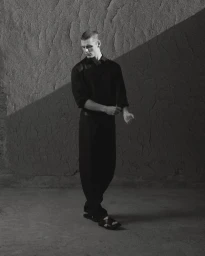 Monochrome fashion portrait of a male model in a black shirt and trousers against a textured stone wall.