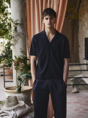 Portrait of a male model wearing a navy polo shirt and matching pants. The setting is a villa with neutral drapes.