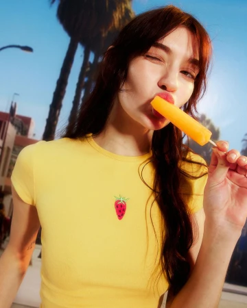 Young woman in yellow top with strawberry design eating an orange popsicle against a backdrop of palm trees and a bright blue sky.
