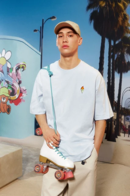 Portrait of a young adult male wearing a light blue t-shirt and baseball cap, holding roller skates with a graphic wall backdrop.