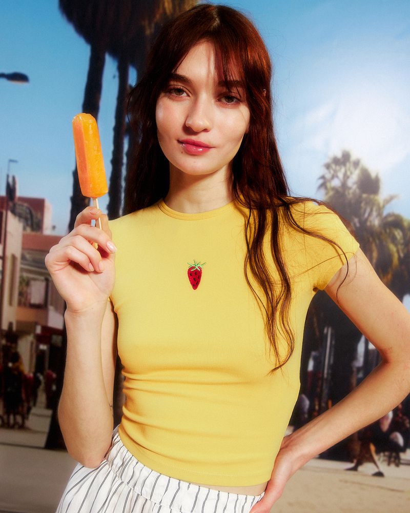 Young woman in a yellow t-shirt with a strawberry emblem, holding a yellow popsicle outdoors in a tropical-themed urban setting.