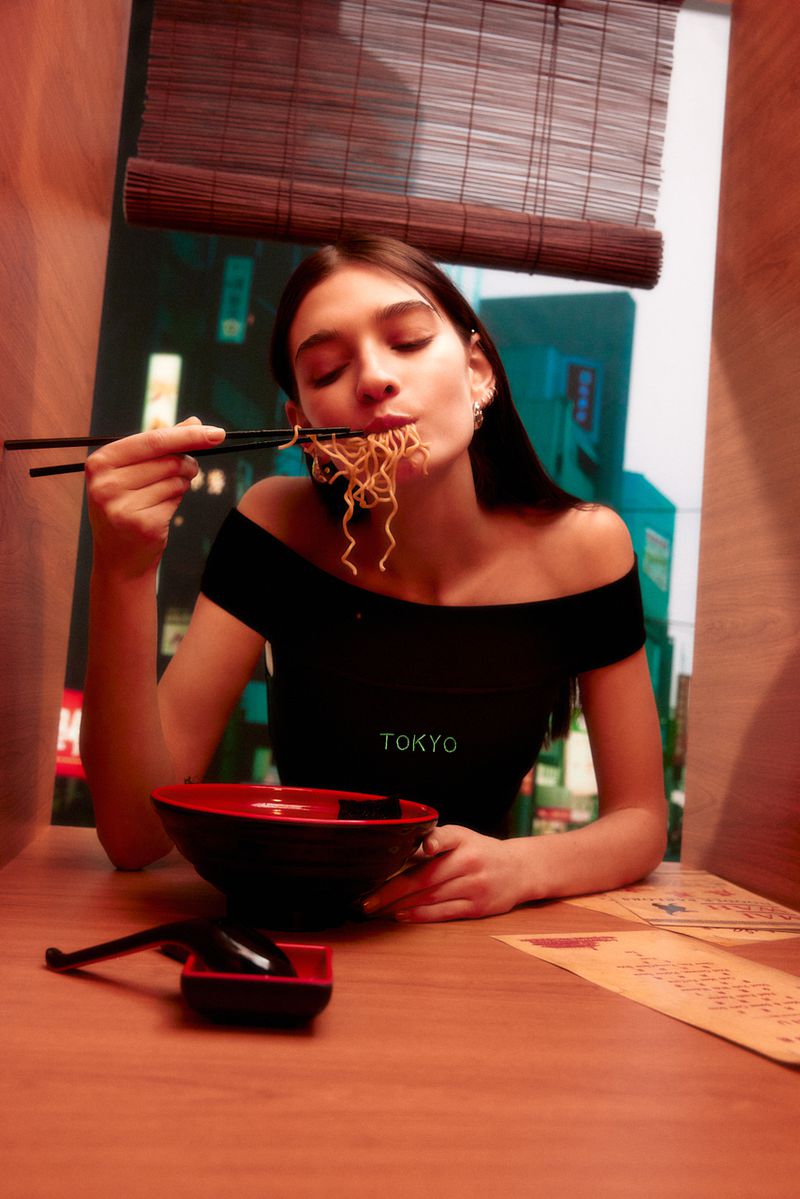 A woman with long brown hair is eating noodles with chopsticks at a table in a booth, with a bamboo blind in the background.