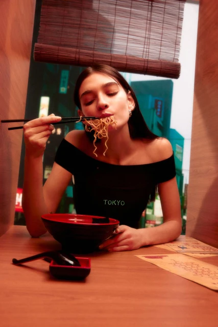 A woman with long brown hair is eating noodles with chopsticks at a table in a booth, with a bamboo blind in the background.