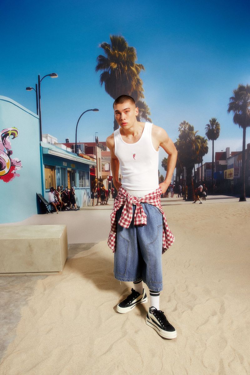 Young adult male in white tank top, denim shorts, and sneakers poses on beach boardwalk backdrop with palm trees and street scene.