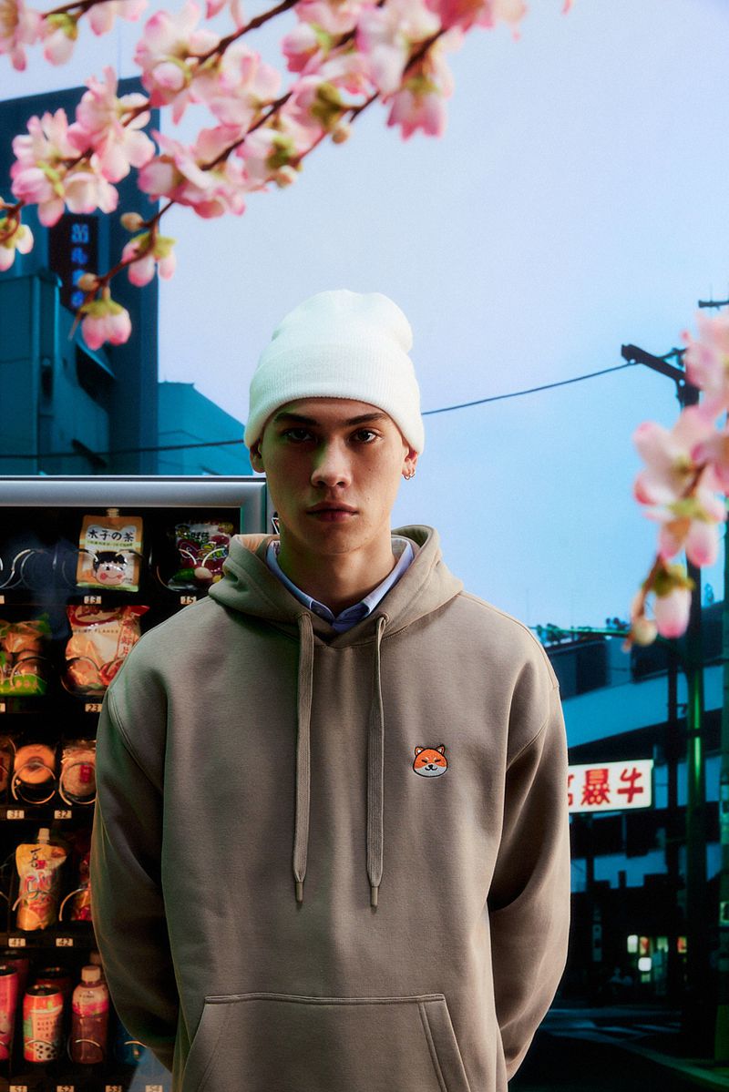 Portrait of a young adult male wearing a hoodie and beanie, standing in front of a Japanese vending machine with blooming cherry blossoms.