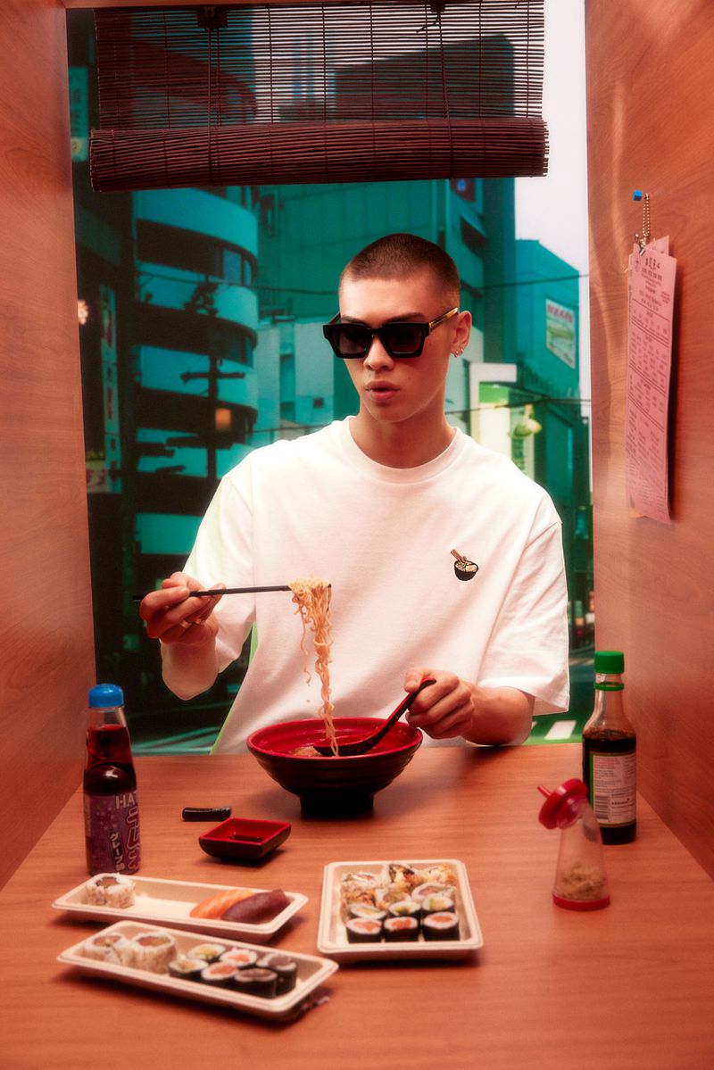 Indoor shot of a young adult male model in sunglasses eating noodles at a table with sushi, against an out-of-focus city background.