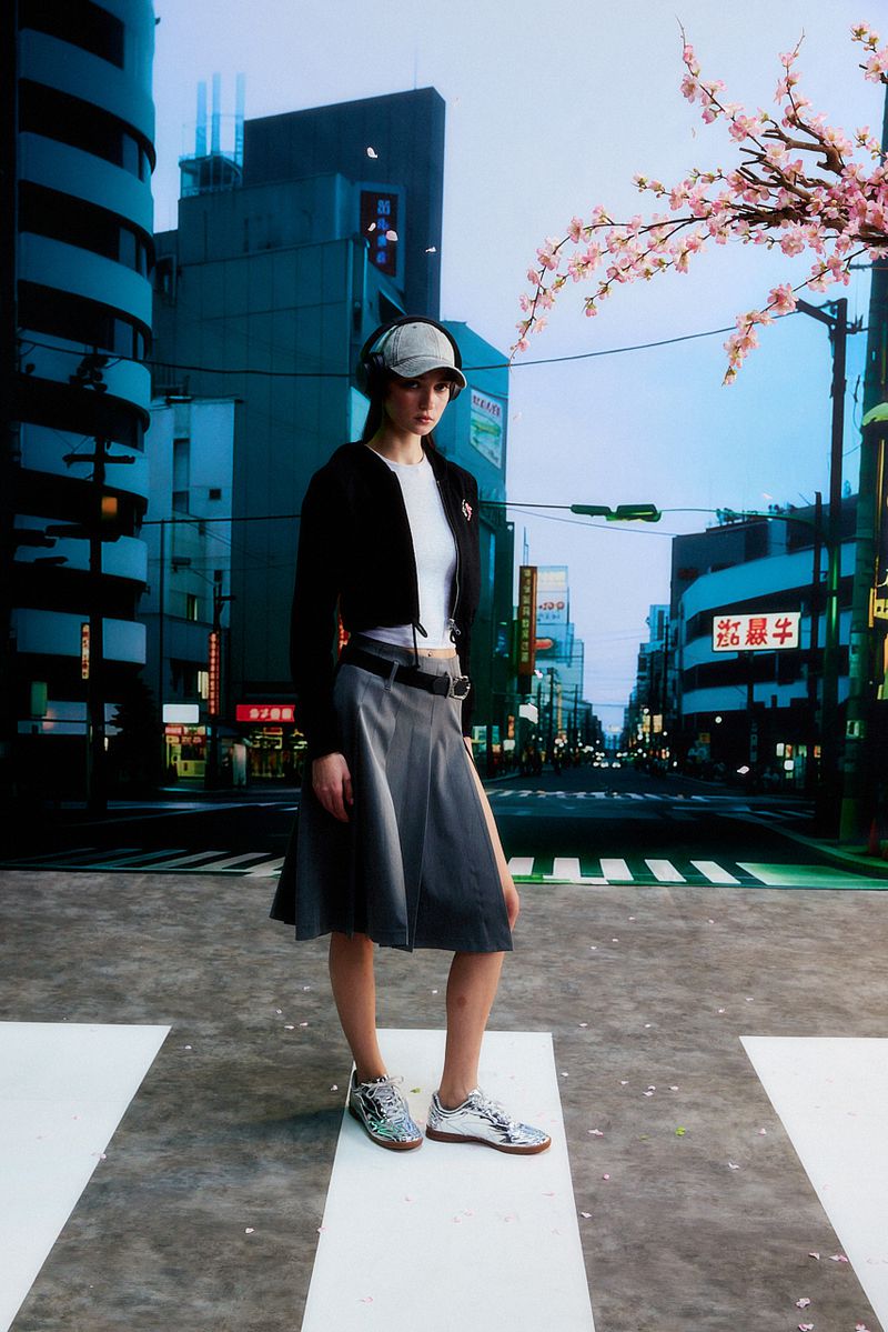 A full shot of a young woman posing on a crosswalk in a stylish outfit against an urban backdrop with cherry blossoms.