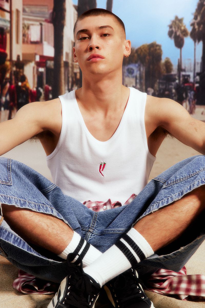 A young adult sits cross-legged in casual streetwear: a white tank top, baggy jeans, striped socks, and sneakers, against a blurred street.