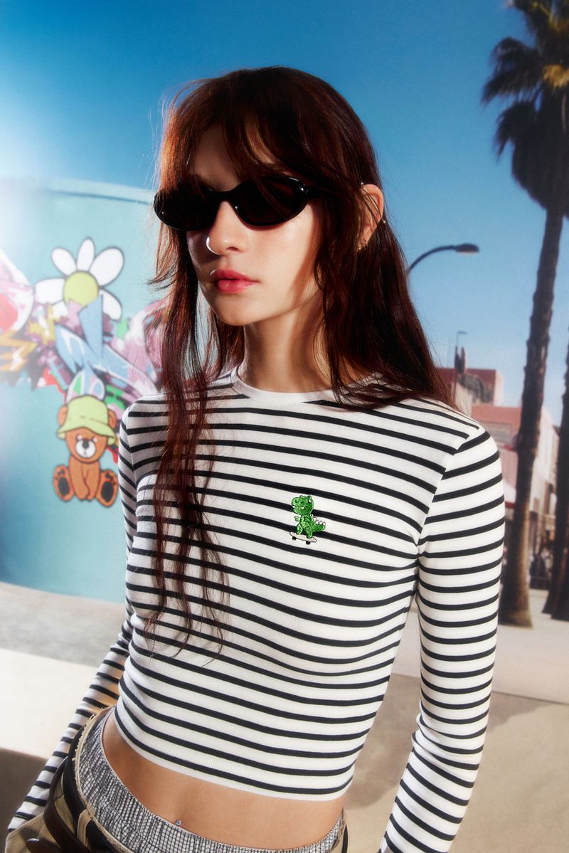 A young woman with long brown hair poses in a striped long-sleeve shirt, sunglasses, and casual pants against a stylized outdoor backdrop.
