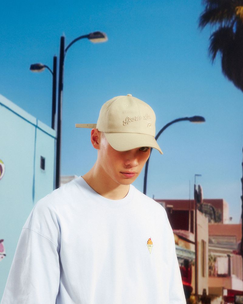 Man in a light blue t-shirt and tan baseball cap poses in an urban setting with palm trees and blue skies in the background on a sunny day.