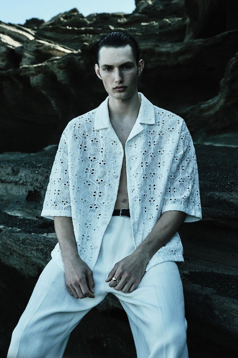 A young adult man poses outdoors, wearing a patterned white shirt and pants, with a dark belt, against a textured rock background.