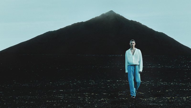 A male fashion model in a white shirt and blue pants walks against a stark landscape backdrop with a dark, imposing mountain.