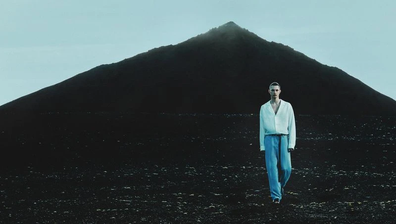 A male fashion model in a white shirt and blue pants walks against a stark landscape backdrop with a dark, imposing mountain.