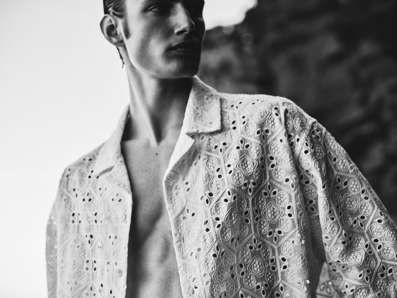 Monochrome portrait of a young adult man wearing an open, patterned, collared shirt, partially revealing his chest against a blurred rock backdrop.