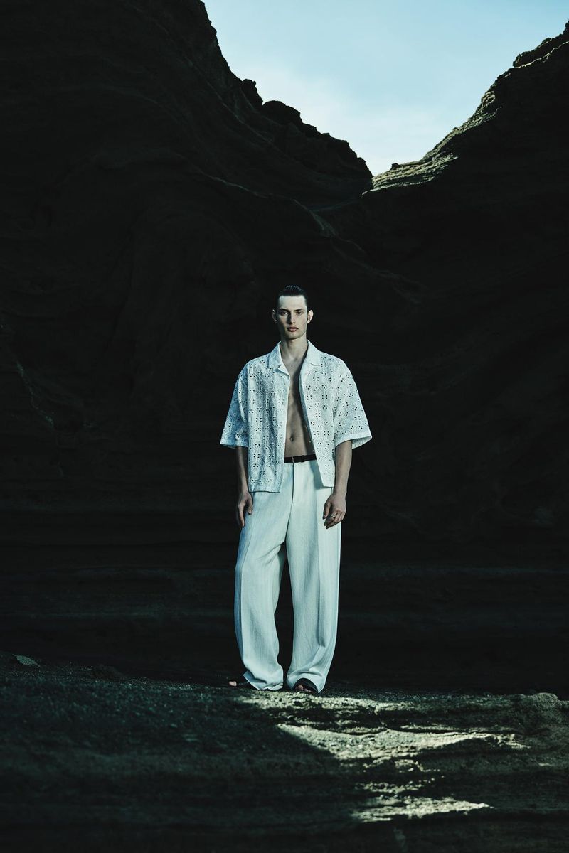 A stylish young man wearing a white shirt and trousers stands against a dark, rocky backdrop with a hint of bright sky above.