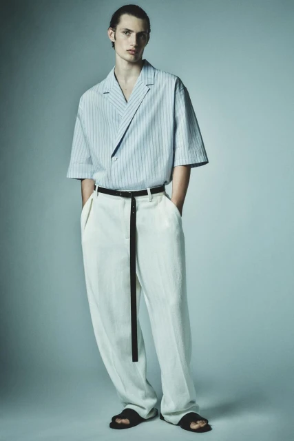 A young adult male fashion model poses in a blue and white striped shirt, white pants, black belt, and sandals against a grey background.