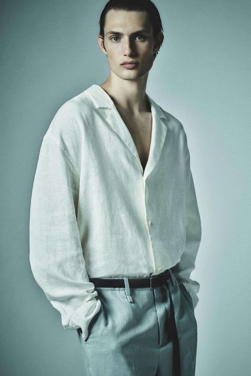 A young adult male poses in a white linen shirt and light blue trousers in a studio shot with soft lighting and a neutral background.