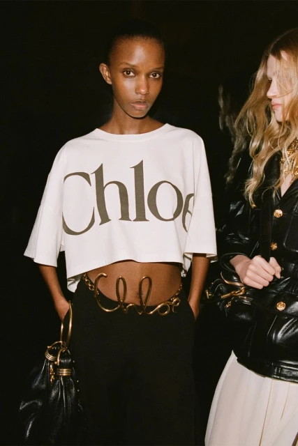 Two women in a dark setting. One woman with an olive complexion wears a cropped shirt with 'Chloe' and a 'Chloe' belt.