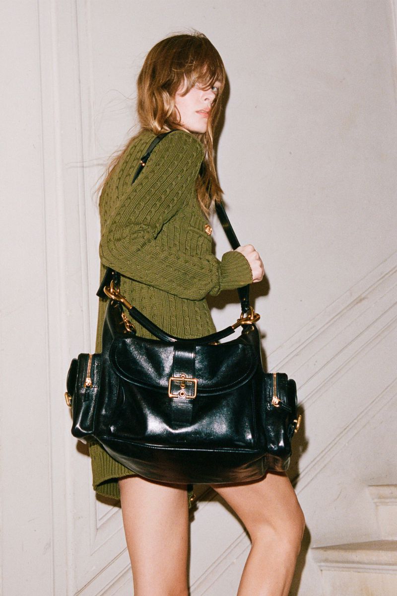 A woman wearing a green cable knit sweater dress and carrying a black leather satchel bag is walking up stairs inside.