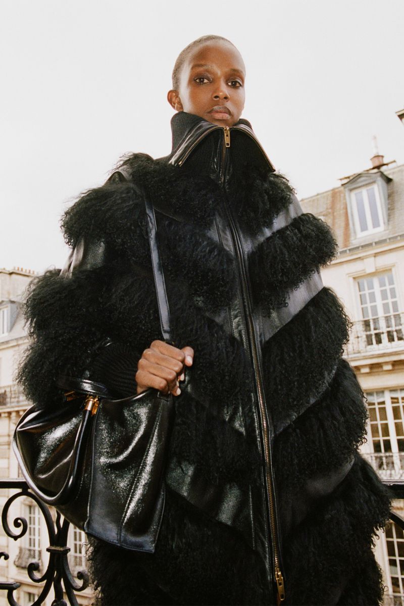 A woman stands outside wearing a textured black coat and carrying a glossy black handbag, with a neutral expression, and buildings in background.