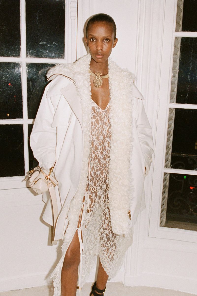 Fashion model wearing an off-white coat and lacy dress poses indoors by a window for a fashion editorial, showing modern style.