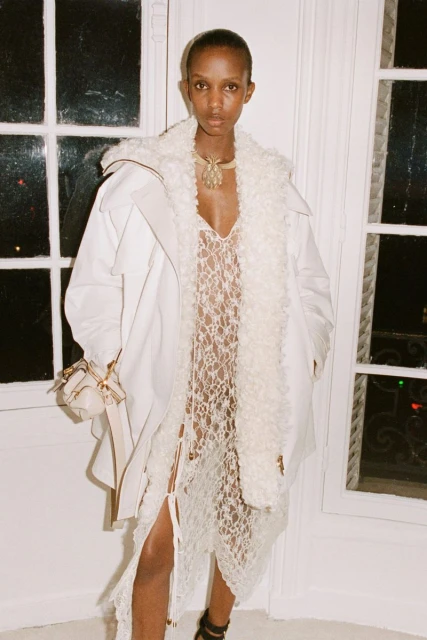 Fashion model wearing an off-white coat and lacy dress poses indoors by a window for a fashion editorial, showing modern style.