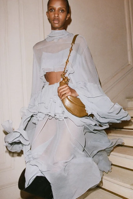 Elegant Black woman in a flowing grey dress and gold purse, walking down stairs against an off-white wall, fashion shot.