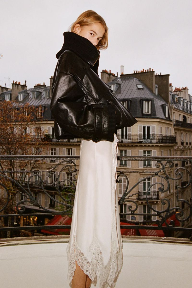 Woman in black leather jacket and white lace-trimmed skirt poses against a Paris balcony with city skyline backdrop on overcast day.