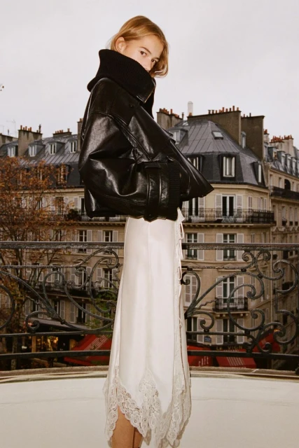 Woman in black leather jacket and white lace-trimmed skirt poses against a Paris balcony with city skyline backdrop on overcast day.