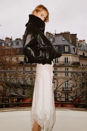 Woman in black leather jacket and white lace-trimmed skirt poses against a Paris balcony with city skyline backdrop on overcast day.