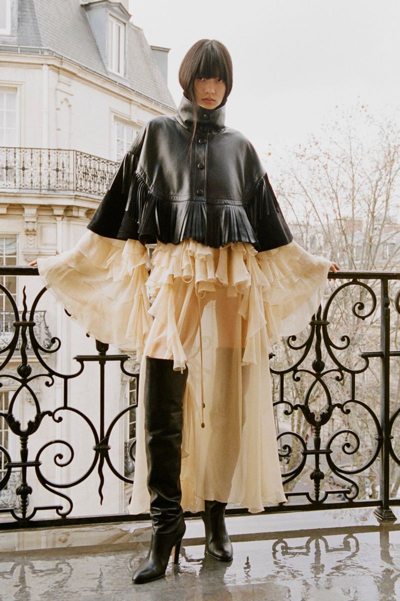 Woman in a black jacket and flowing dress stands on a balcony with wrought iron railings and a city building in the background.