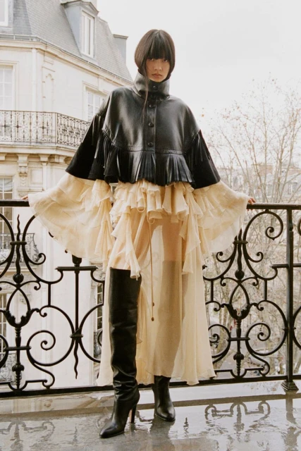 Woman in a black jacket and flowing dress stands on a balcony with wrought iron railings and a city building in the background.