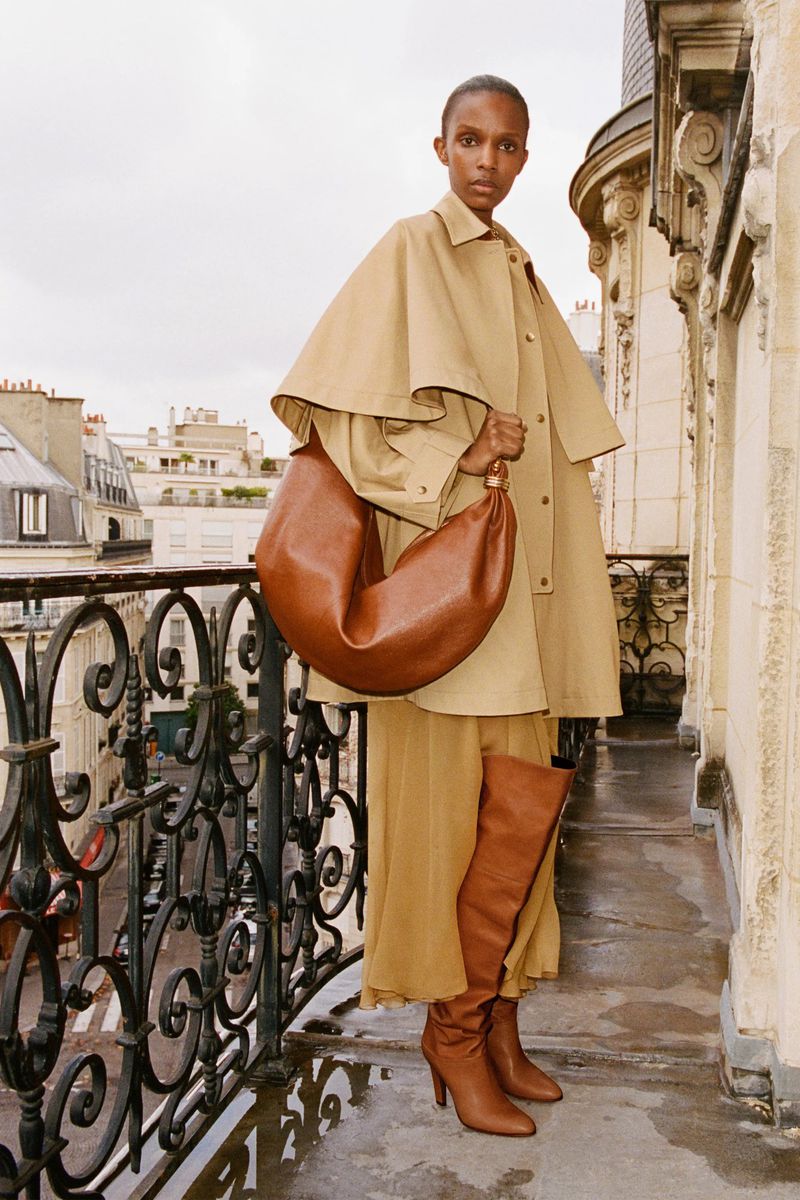 An adult woman poses on a balcony wearing a tan trench coat, brown leather boots, and a brown leather hobo bag in an urban setting.