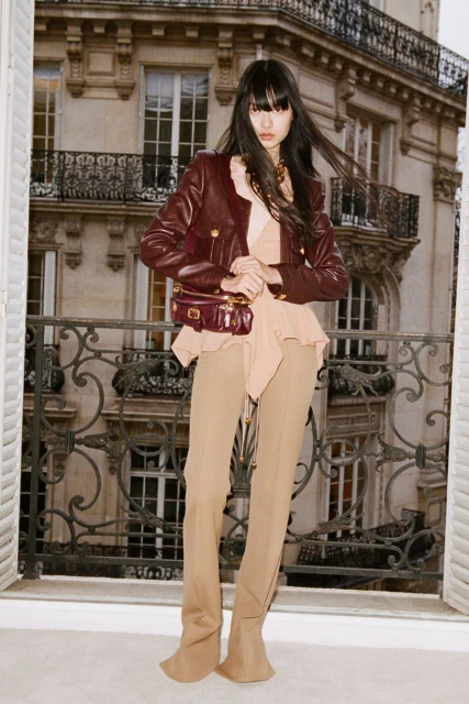 Brunette woman in a leather jacket and beige pants poses on a balcony with an ornamental iron railing in front of an ornate building.