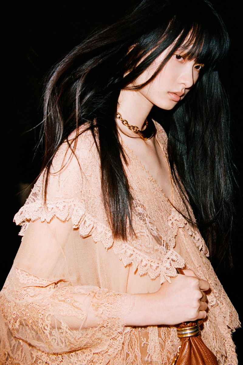 A woman with long dark hair wears a peach-colored lace dress and gold necklace against a dark backdrop, holding a brown leather purse.