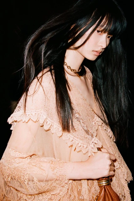 A woman with long dark hair wears a peach-colored lace dress and gold necklace against a dark backdrop, holding a brown leather purse.