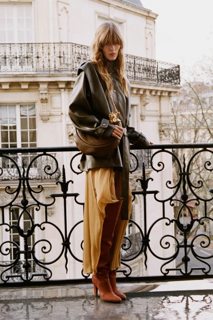 A woman standing on a balcony wearing a brown leather jacket, tan dress and brown boots, with a cityscape in the background.